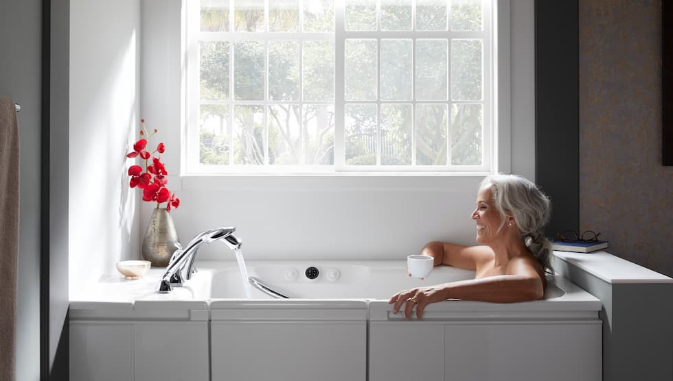 An elderly woman comfortably taking a bath in a walk-in tub with an open door, looking out a window.