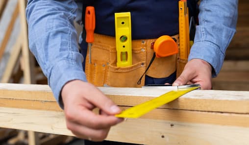A man stands with a wooden plank and tools in hand, focused on his woodworking project.